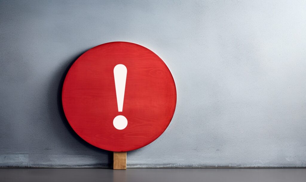 グレーの壁を背景にした、赤い標識上の感嘆符（びっくりマーク）。「注意」や「警告」を意味するイメージ